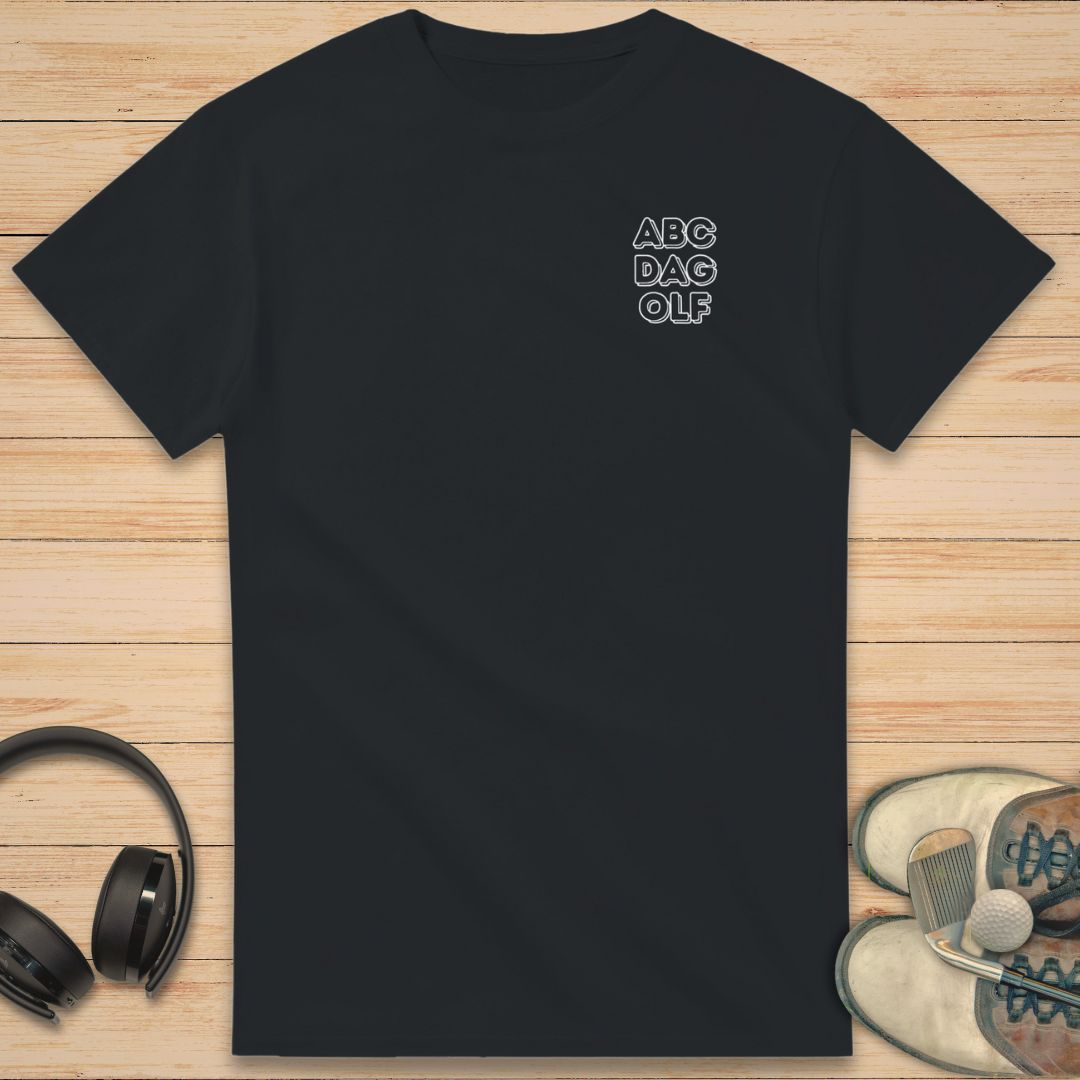 ABC Da Golf T-Shirt in Black on a light wooden floor lying next to headphones and golf shoes.