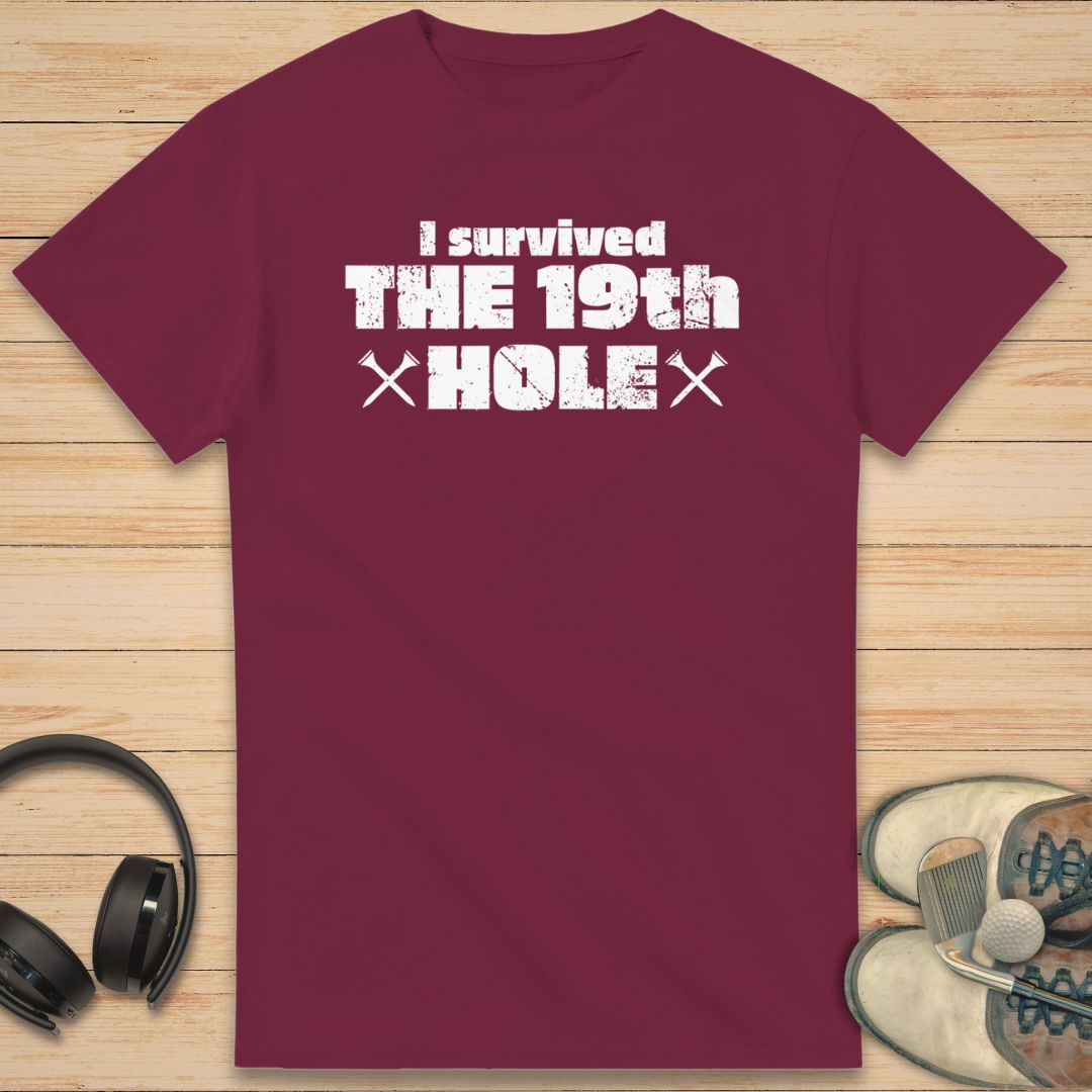 The 19th Hole T-Shirt in Maroon on a light wooden floor lying next to headphones and golf shoes.