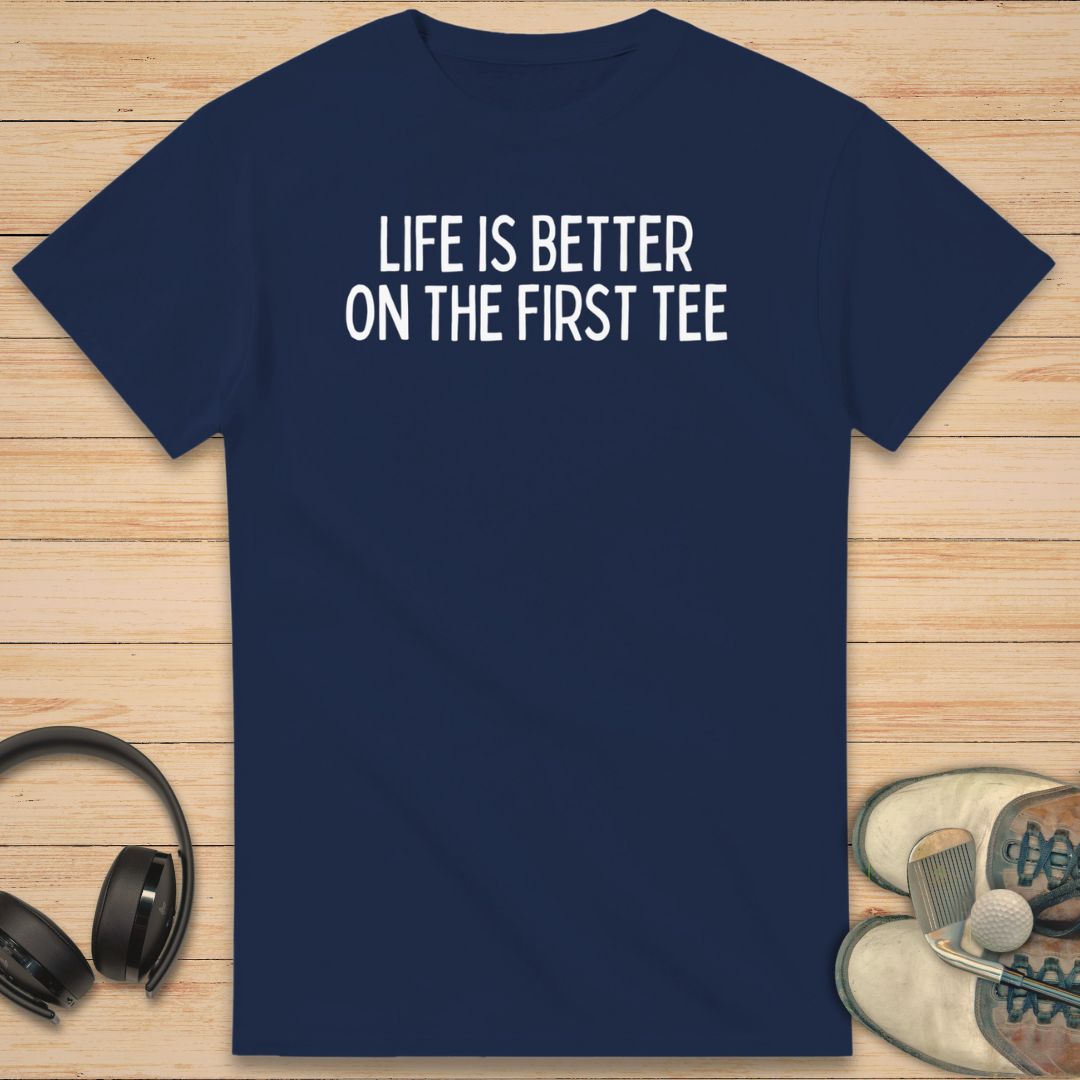 First Tee T-Shirt in Navy on a light wooden floor lying next to headphones and golf shoes.