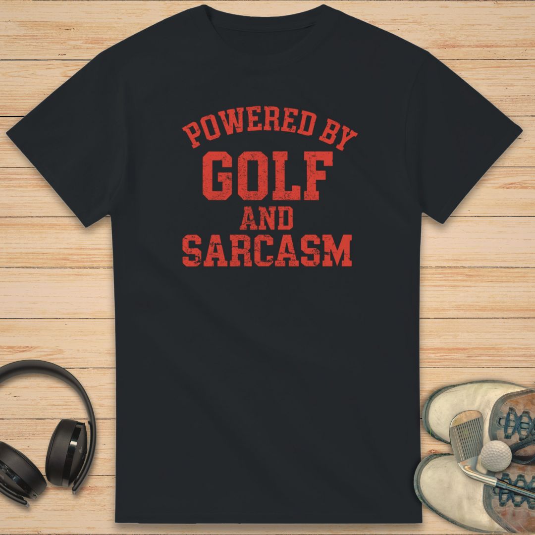Powered By T-Shirt in Black on a light wooden floor lying next to headphones and golf shoes.