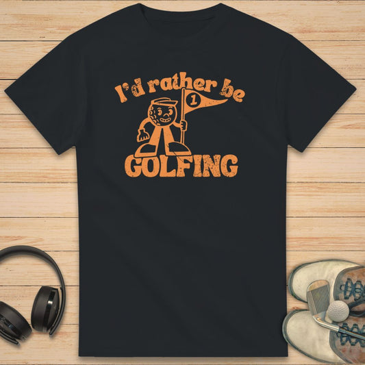 Be Golfing Mascot T-Shirt in Black on a light wooden floor lying next to headphones and golf shoes.