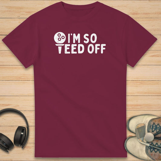 Teed Off T-Shirt in Maroon on a light wooden floor lying next to headphones and golf shoes.