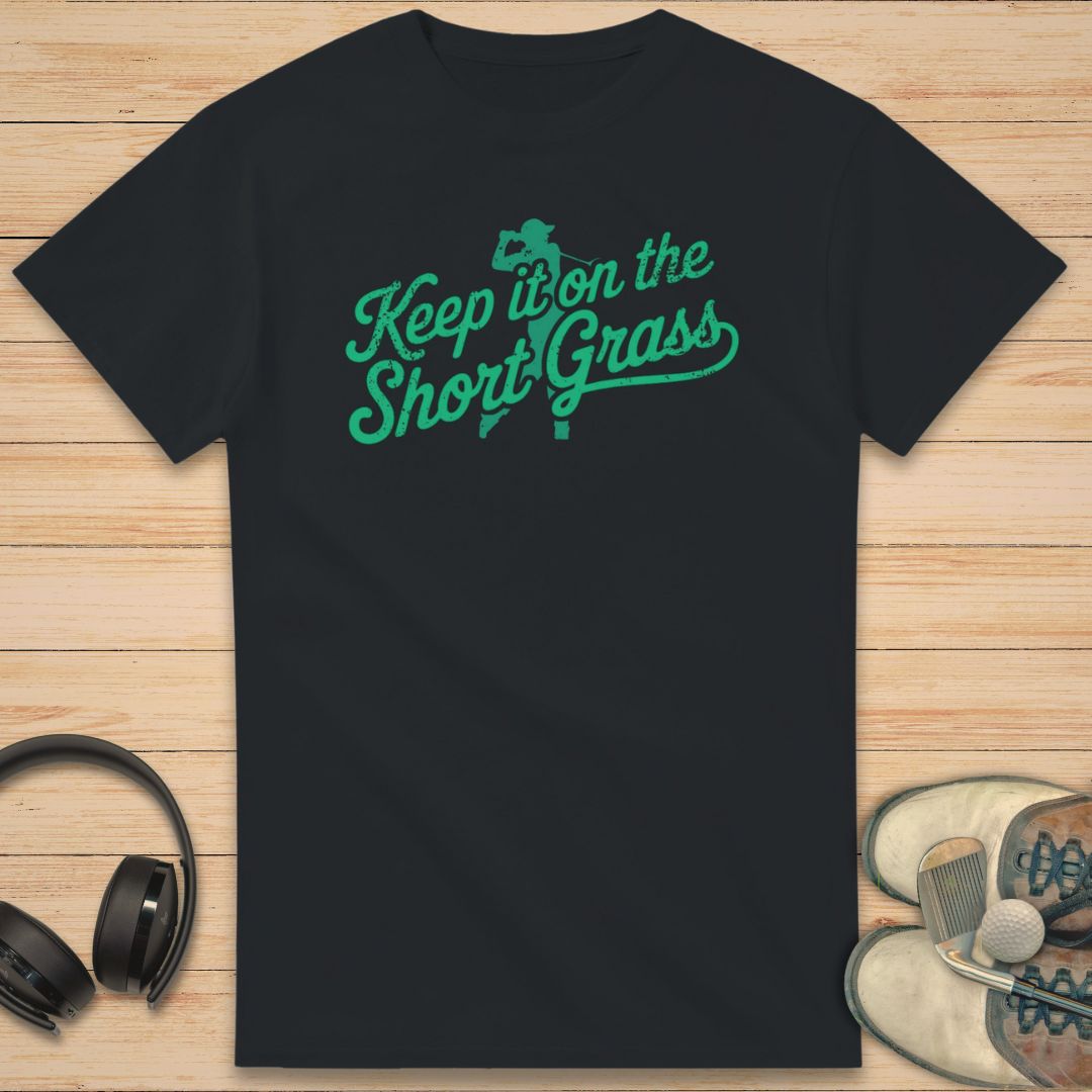 Black t-shirt with green text 'Keep it on the Short Grass' on a wooden surface with headphones and shoes.
