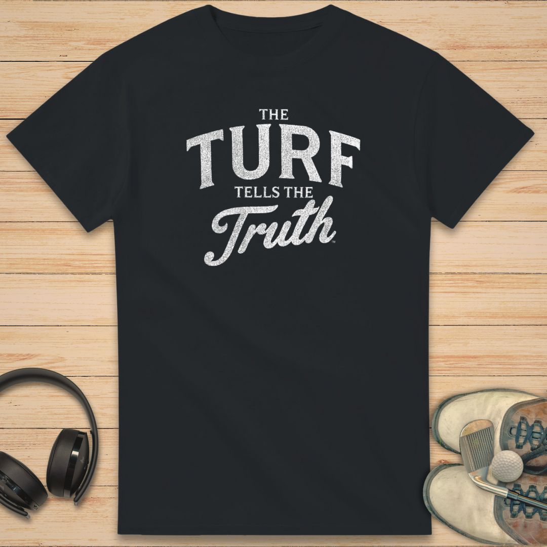 The Turf T-Shirt in Black on a light wooden floor lying next to headphones and golf shoes.