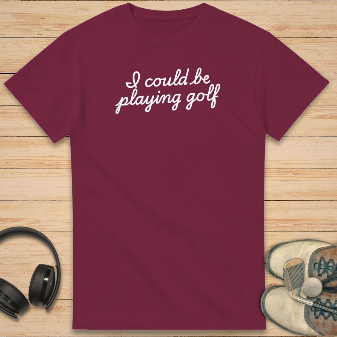 Could Be T-Shirt in Maroon on a light wooden floor lying next to headphones and golf shoes.