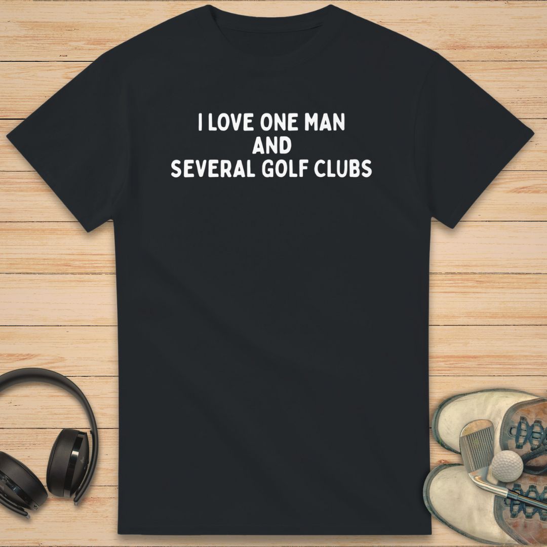 One Man T-Shirt in Black on a light wooden floor lying next to headphones and golf shoes.