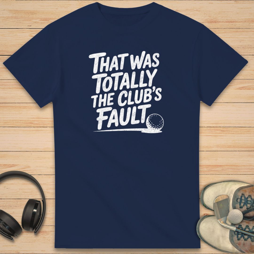 Club's Fault T-Shirt in on a light wooden floor lying next to headphones and golf shoes.