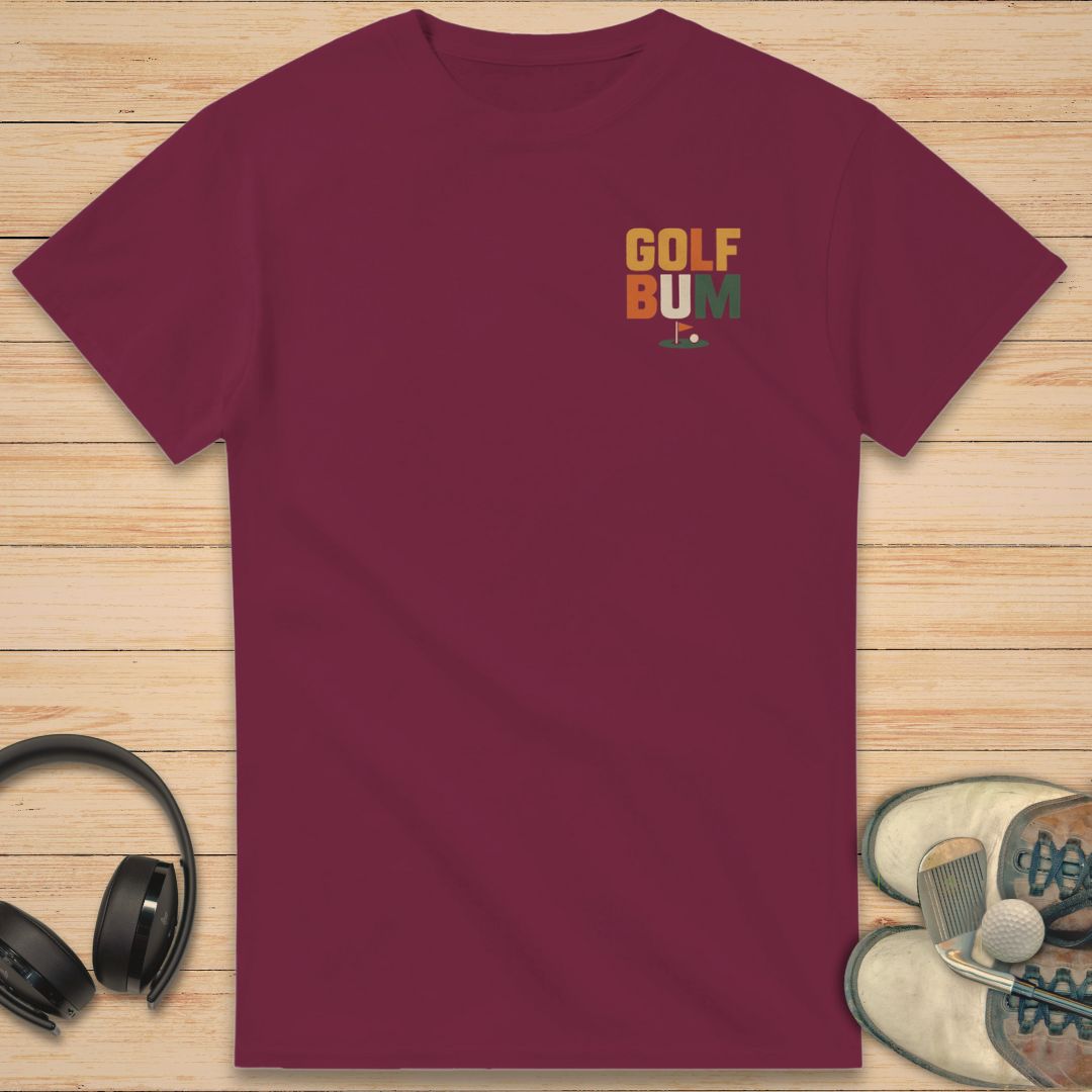 Golf Bum T-Shirt in Maroon on a light wooden floor lying next to headphones and golf shoes.