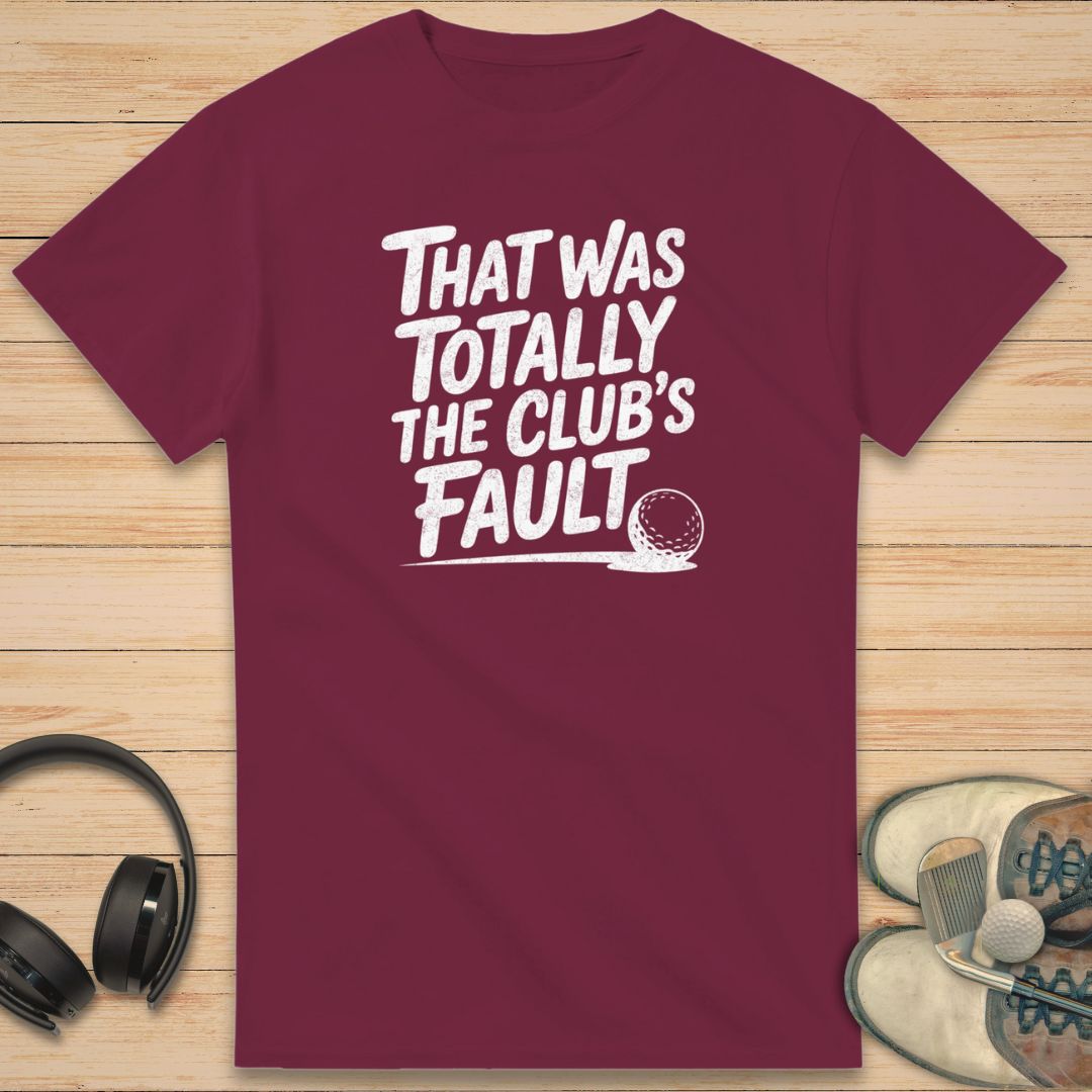 Club's Fault T-Shirt in on a light wooden floor lying next to headphones and golf shoes.