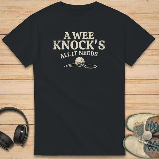 A Wee Knock T-Shirt in Black on a light wooden floor lying next to headphones and golf shoes.