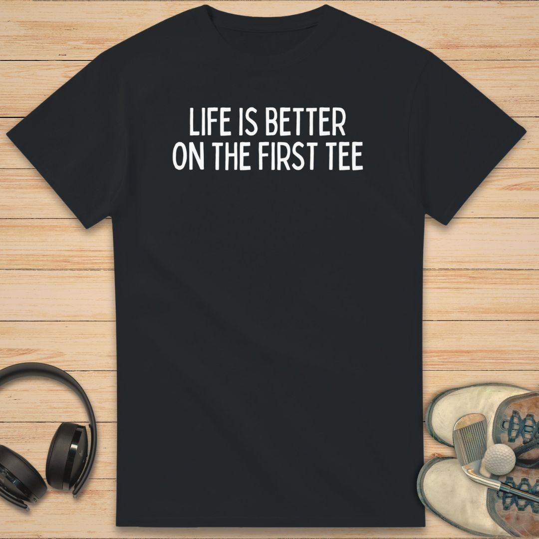 First Tee T-Shirt in Black on a light wooden floor lying next to headphones and golf shoes.