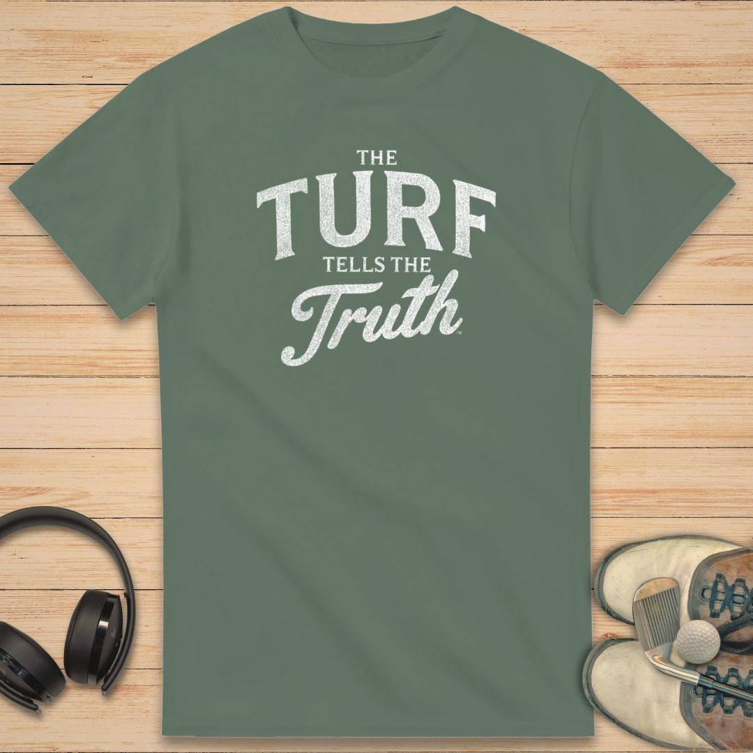 The Turf T-Shirt in Military Green on a light wooden floor lying next to headphones and golf shoes.