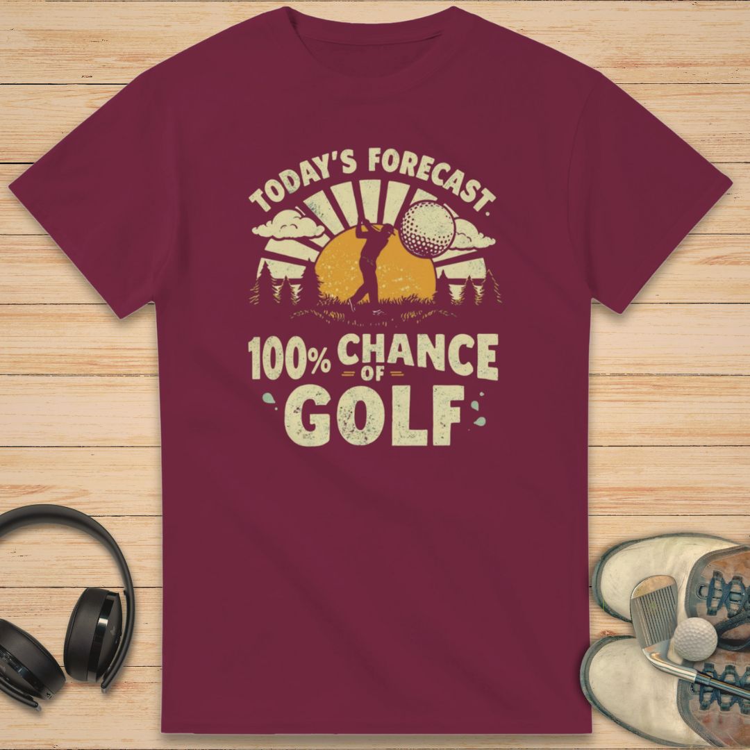 100% Chance of Golf T-Shirt in Maroon on a light wooden floor lying next to headphones and golf shoes.