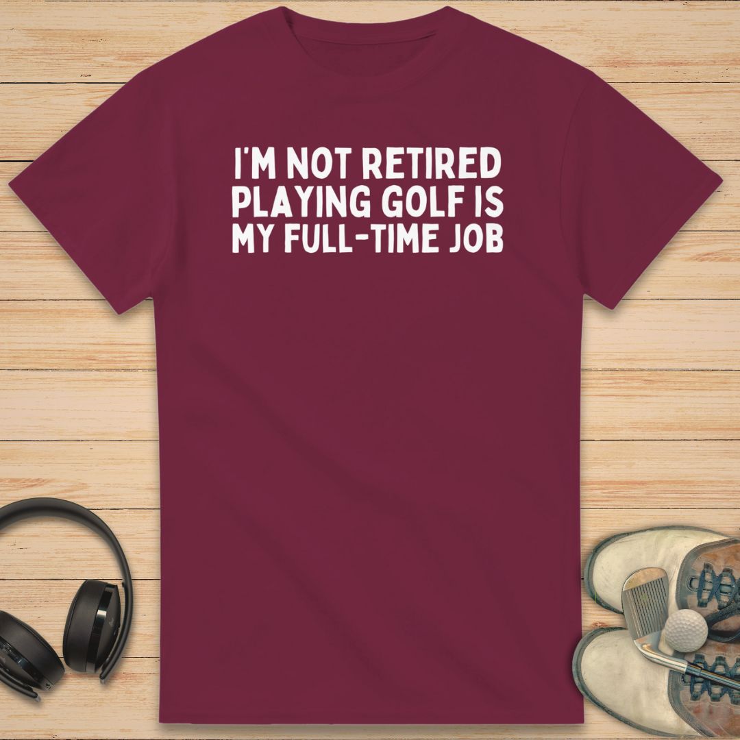 Full-Time Job T-Shirt in Maroon on a light wooden floor lying next to headphones and golf shoes.