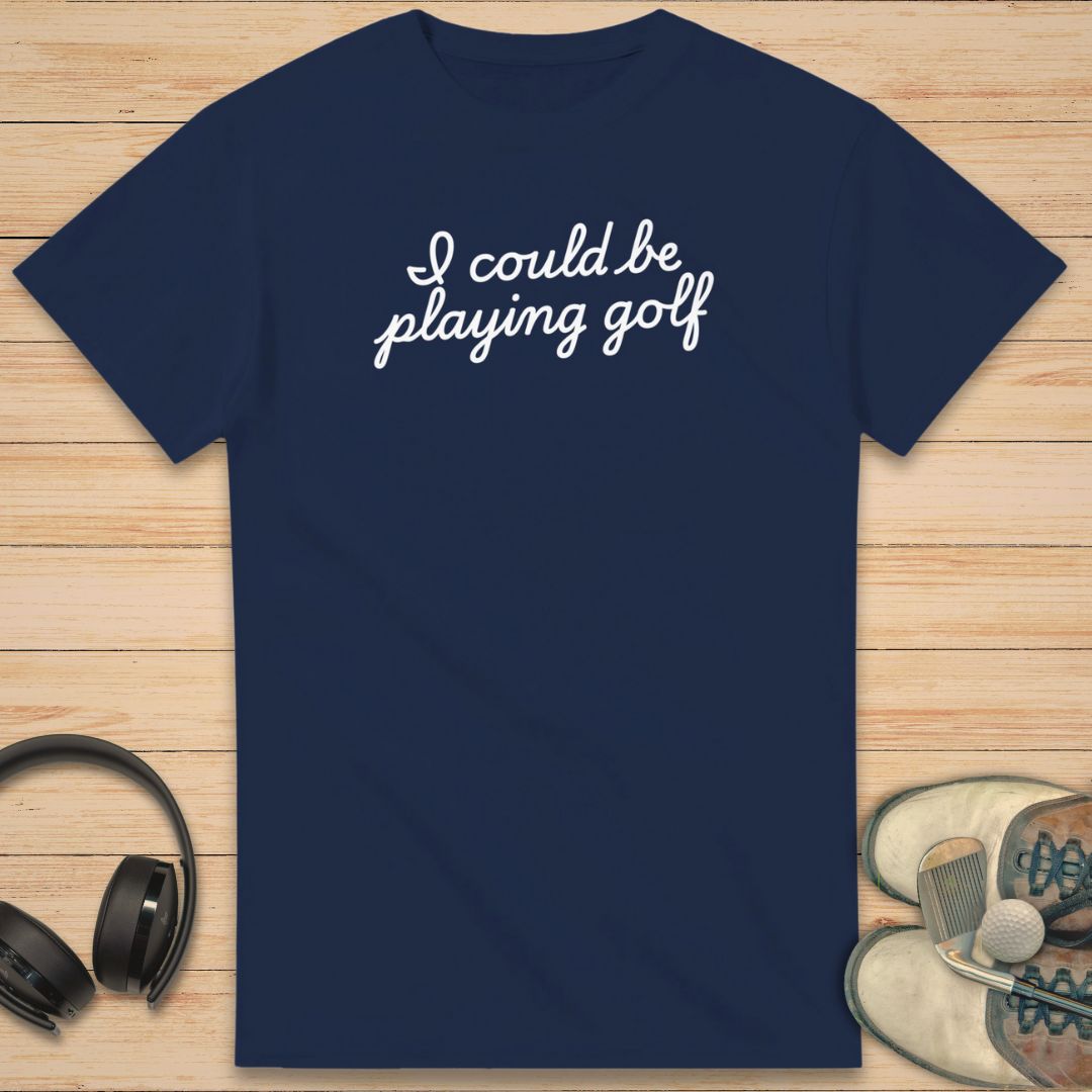 Could Be T-Shirt in Navy on a light wooden floor lying next to headphones and golf shoes.