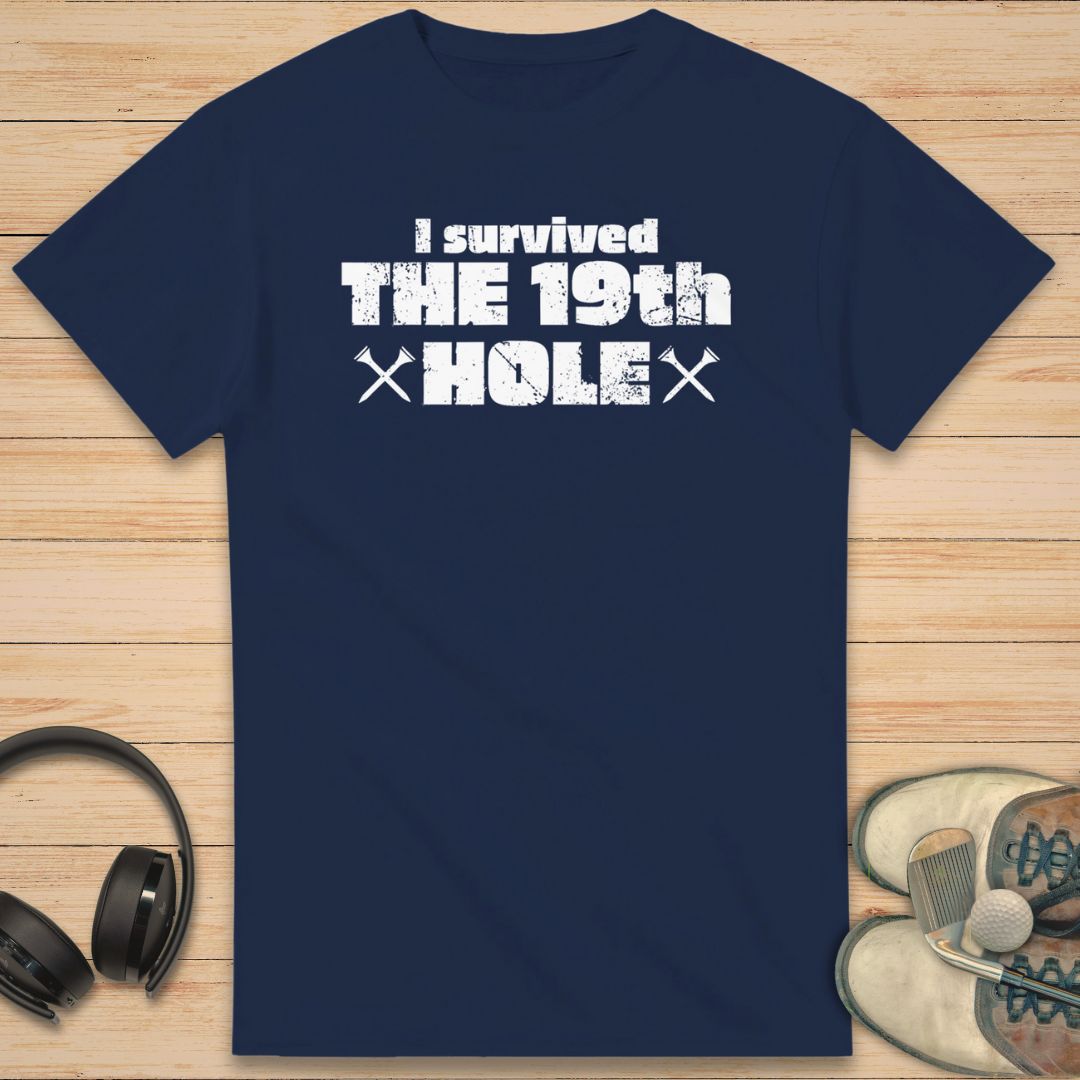 The 19th Hole T-Shirt in Navy on a light wooden floor lying next to headphones and golf shoes.