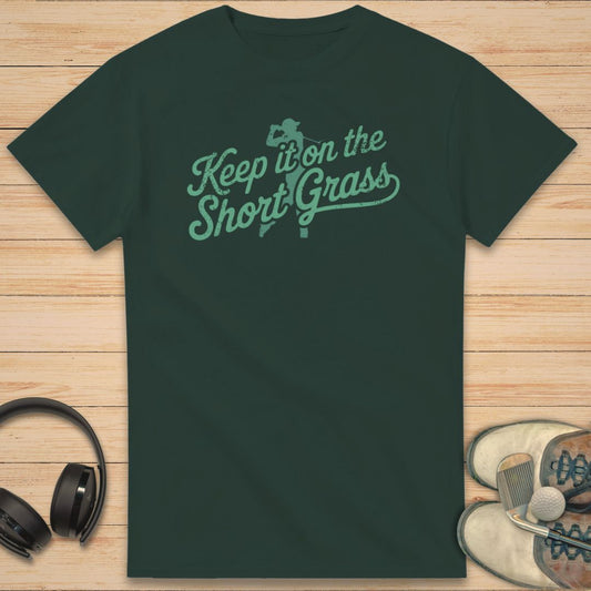 Green t-shirt with 'Keep it on the Short Grass' text on a wooden surface with headphones and shoes.