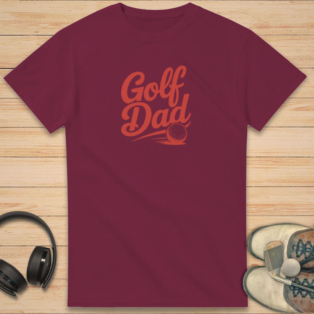 Golf Dad II T-Shirt in Maroon on a light wooden floor lying next to headphones and golf shoes.