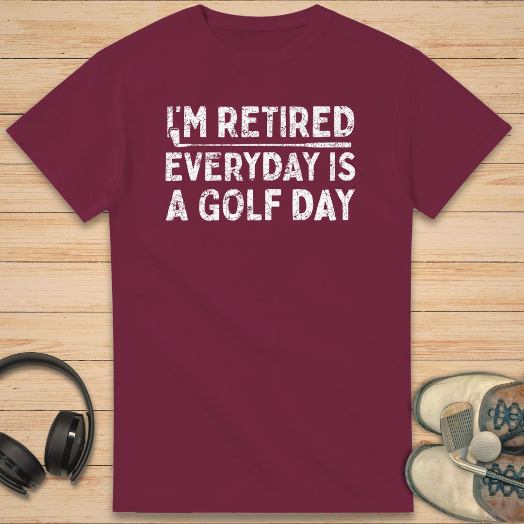 A Golf Day T-Shirt in Maroon on a light wooden floor lying next to headphones and golf shoes.