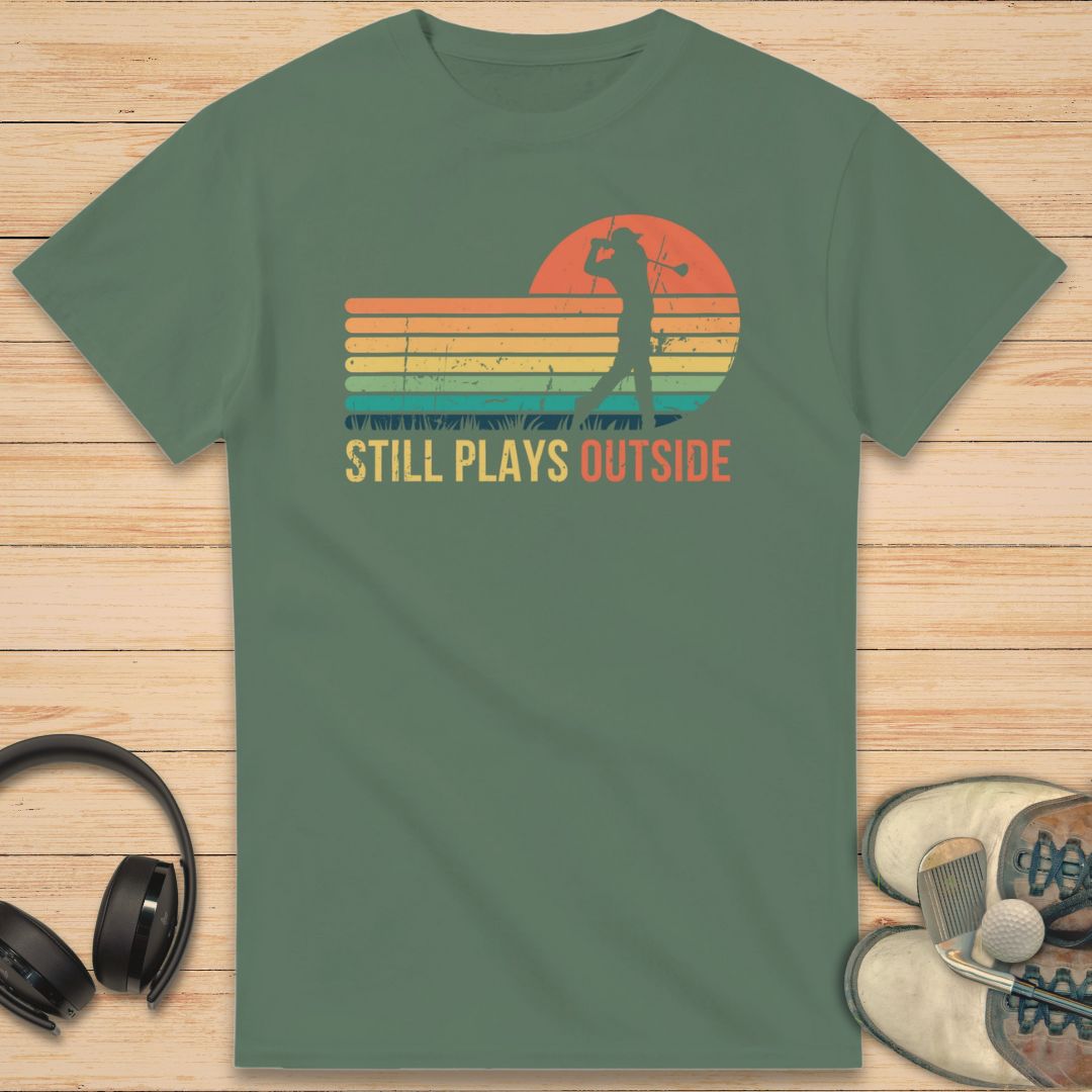 Plays Outside T-Shirt in Military Green on a light wooden floor lying next to headphones and golf shoes.