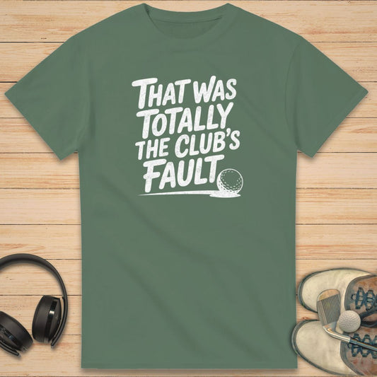Club's Fault T-Shirt in on a light wooden floor lying next to headphones and golf shoes.