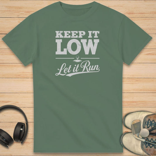 Keep It Low T-Shirt in Military Green on a light wooden floor lying next to headphones and golf shoes.
