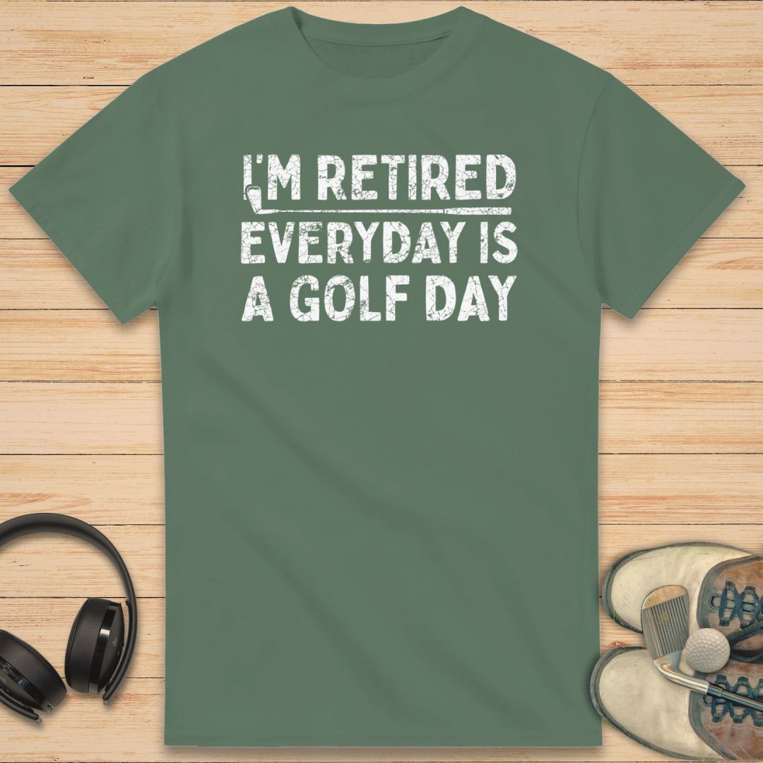 A Golf Day T-Shirt in Military Green on a light wooden floor lying next to headphones and golf shoes.