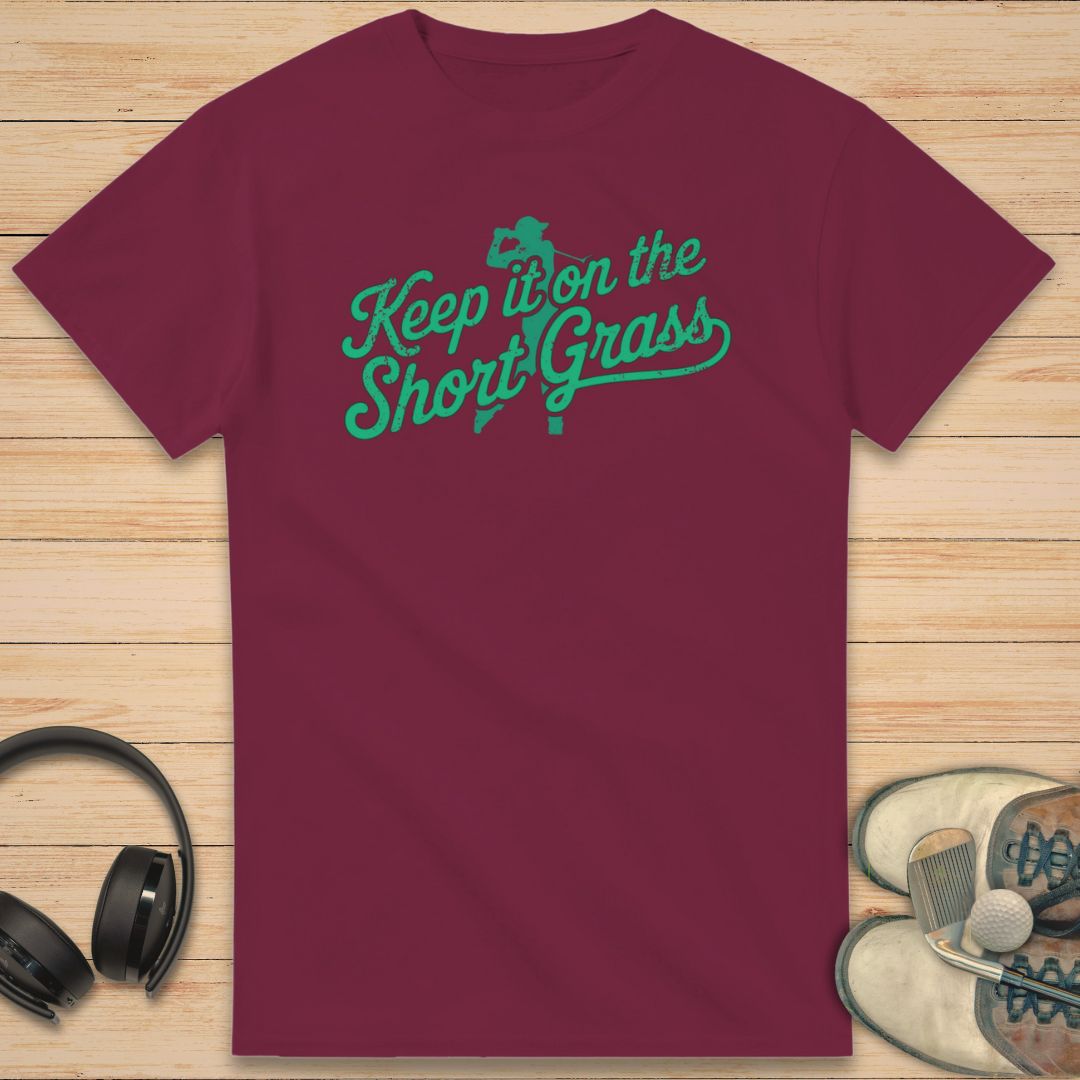 Maroon t-shirt with green text 'Keep it on the Short Grass' on a wooden surface with headphones and shoes.