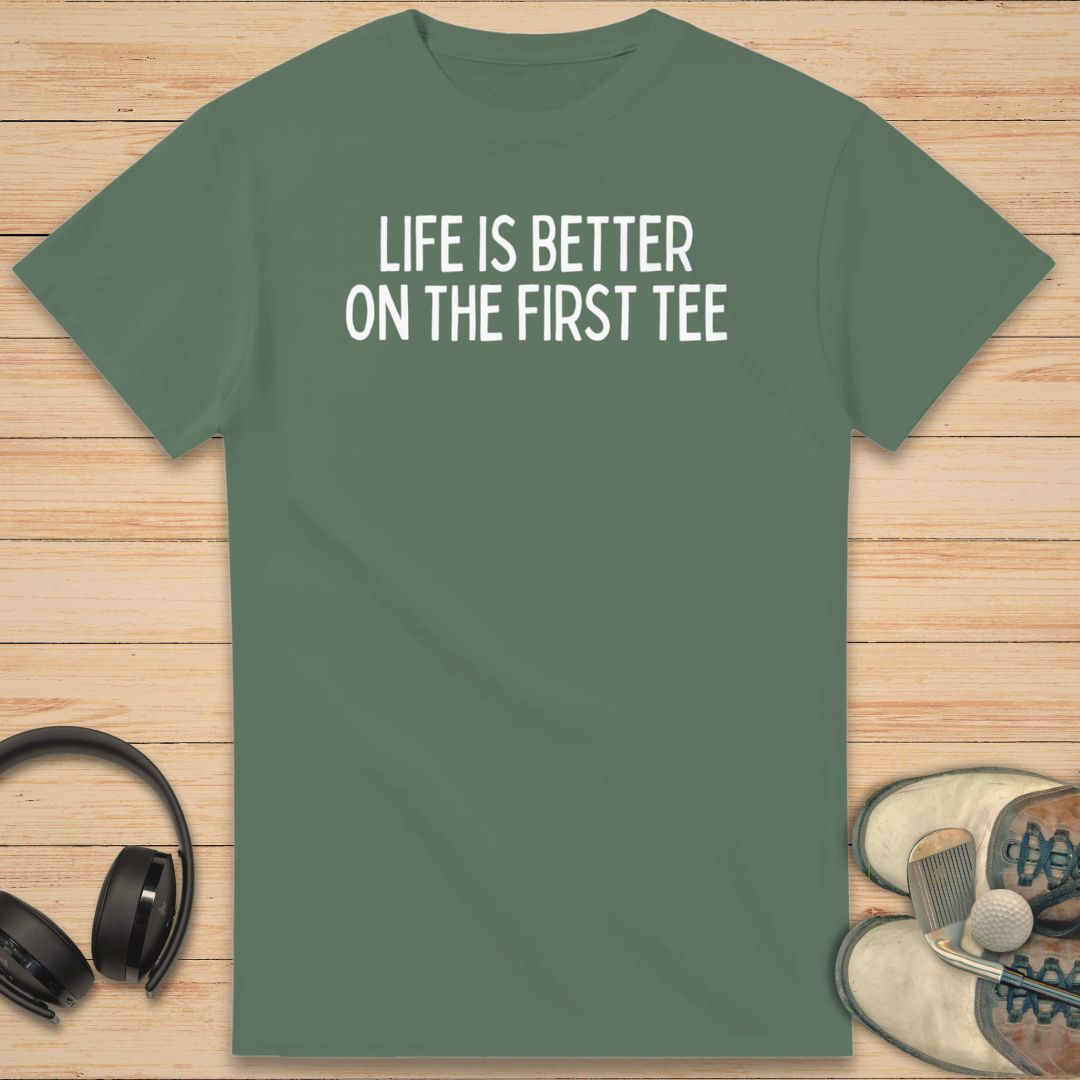 First Tee T-Shirt in Military Green on a light wooden floor lying next to headphones and golf shoes.