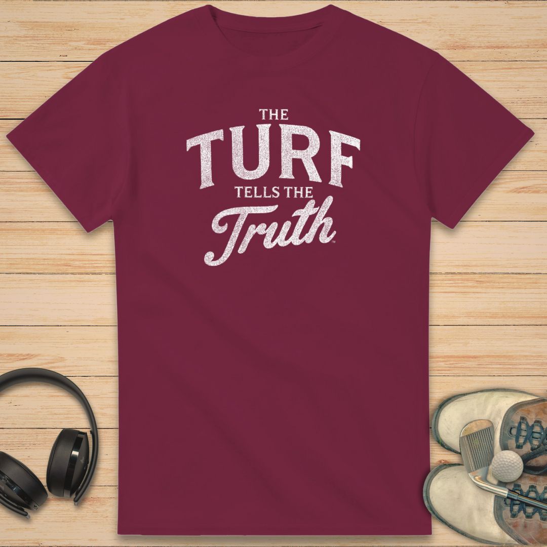 The Turf T-Shirt in Maroon on a light wooden floor lying next to headphones and golf shoes.