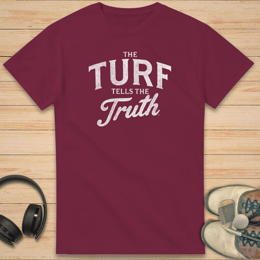The Turf T-Shirt in Maroon on a light wooden floor lying next to headphones and golf shoes.