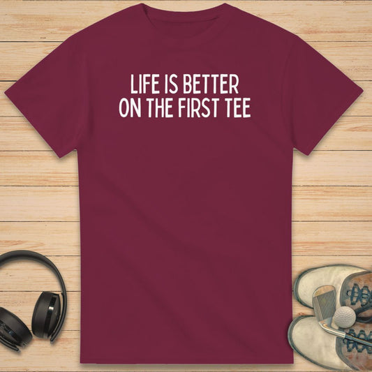 First Tee T-Shirt in Maroon on a light wooden floor lying next to headphones and golf shoes.