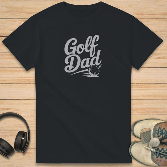 Golf Dad II T-Shirt in Black on a light wooden floor lying next to headphones and golf shoes.