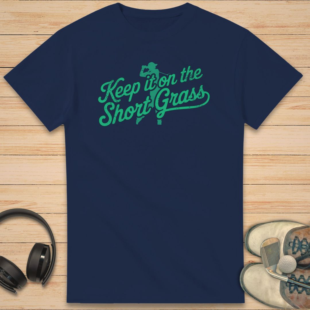 Navy blue t-shirt with green text 'Keep it on the Short Grass' on a wooden surface with headphones and shoes.