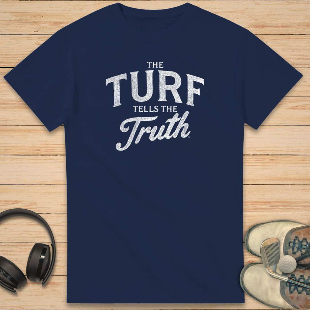 The Turf T-Shirt in Navy on a light wooden floor lying next to headphones and golf shoes.