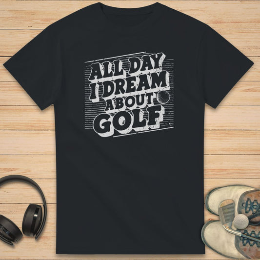 I Dream Golf T-Shirt in Black on a light wooden floor lying next to headphones and golf shoes.