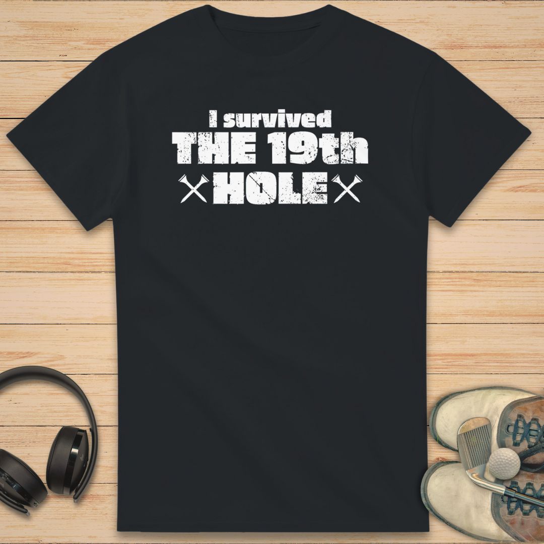 The 19th Hole T-Shirt in Black on a light wooden floor lying next to headphones and golf shoes.