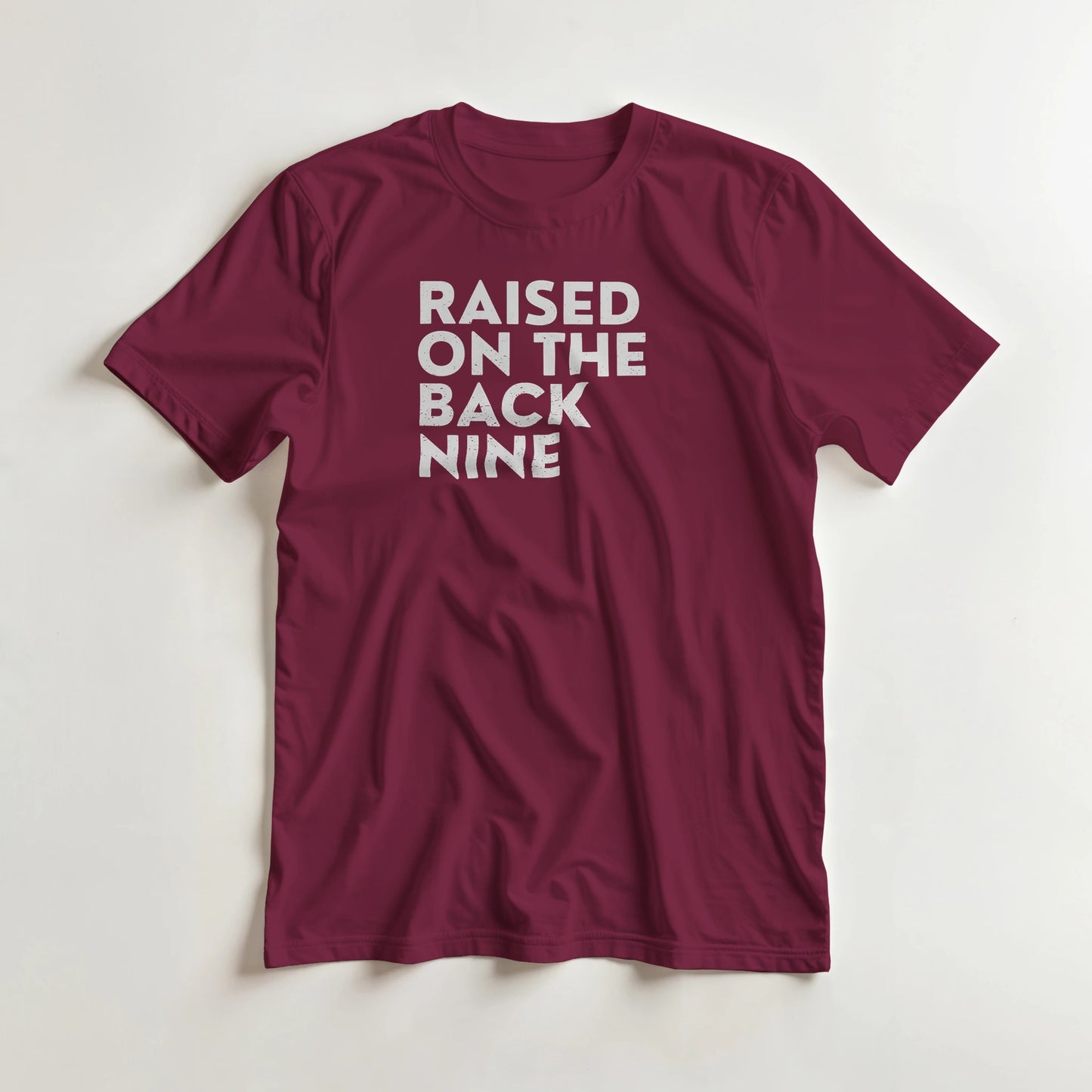 Raised On T-Shirt in Maroon lying on a plain floor. T-shirt is a unisex cut and 100 per cent cotton.