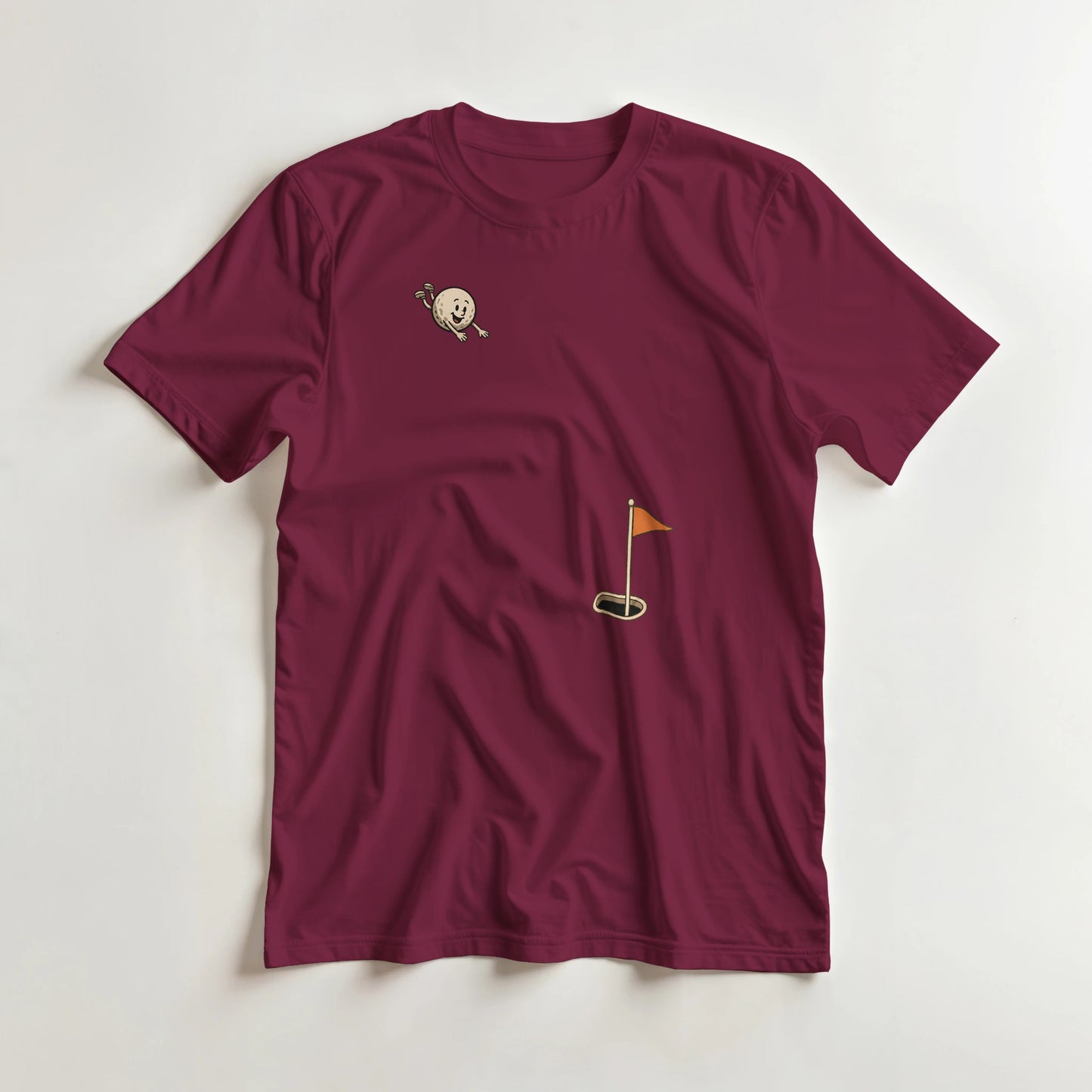 Flying Golf Ball T-Shirt in Maroon lying on a plain floor. T-shirt is a unisex cut and 100 per cent cotton.