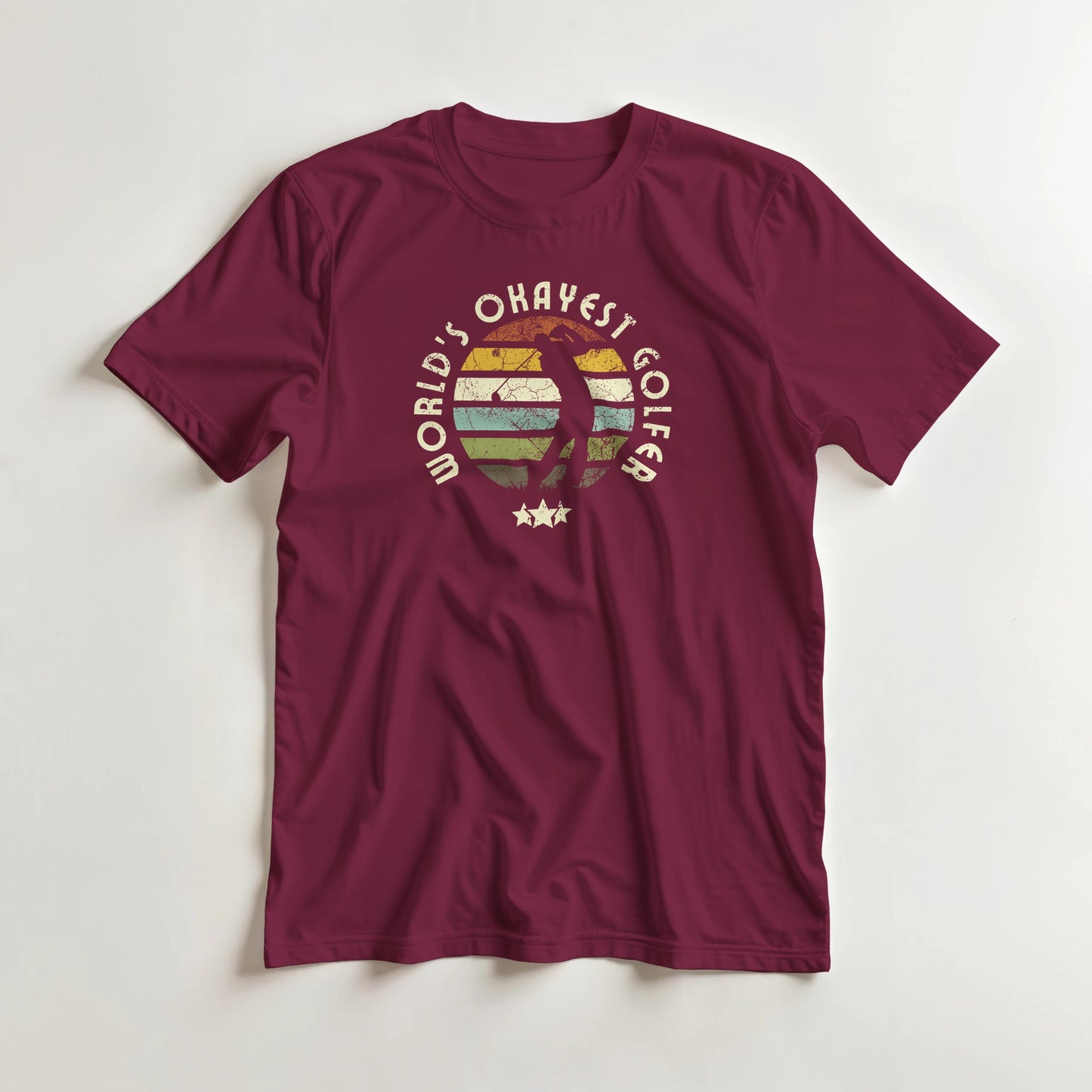 Okayest Golfer T-Shirt in Maroon lying on a plain floor. T-shirt is a unisex cut and 100 per cent cotton.