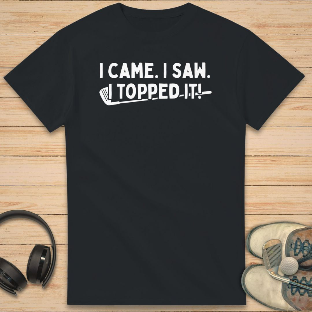 Topped It T-Shirt in Black on a light wooden floor lying next to headphones and golf shoes.