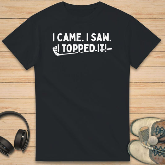 Topped It T-Shirt in Black on a light wooden floor lying next to headphones and golf shoes.
