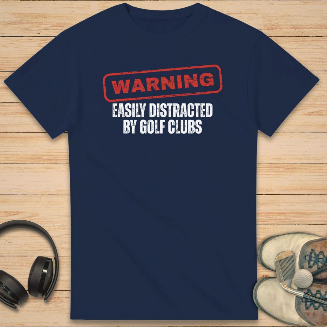 Warning T-Shirt in Navy on a light wooden floor lying next to headphones and golf shoes.