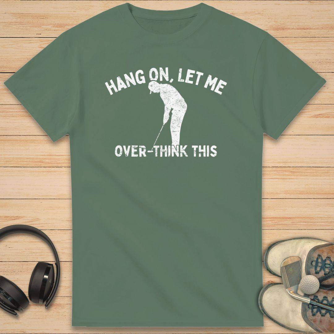 Over-Think T-Shirt in Military Green on a light wooden floor lying next to headphones and golf shoes.