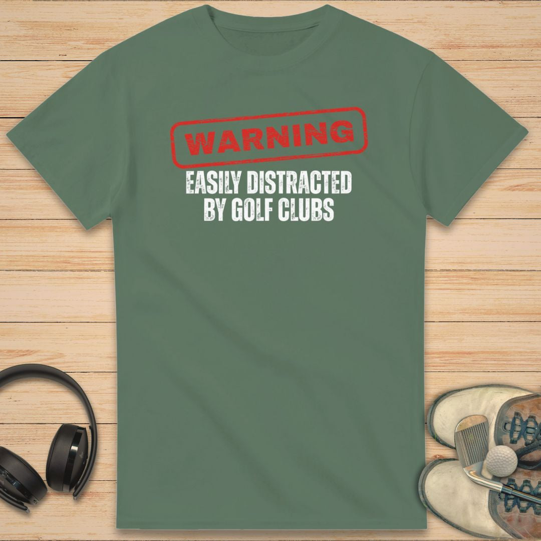 Warning T-Shirt in Military Green on a light wooden floor lying next to headphones and golf shoes.