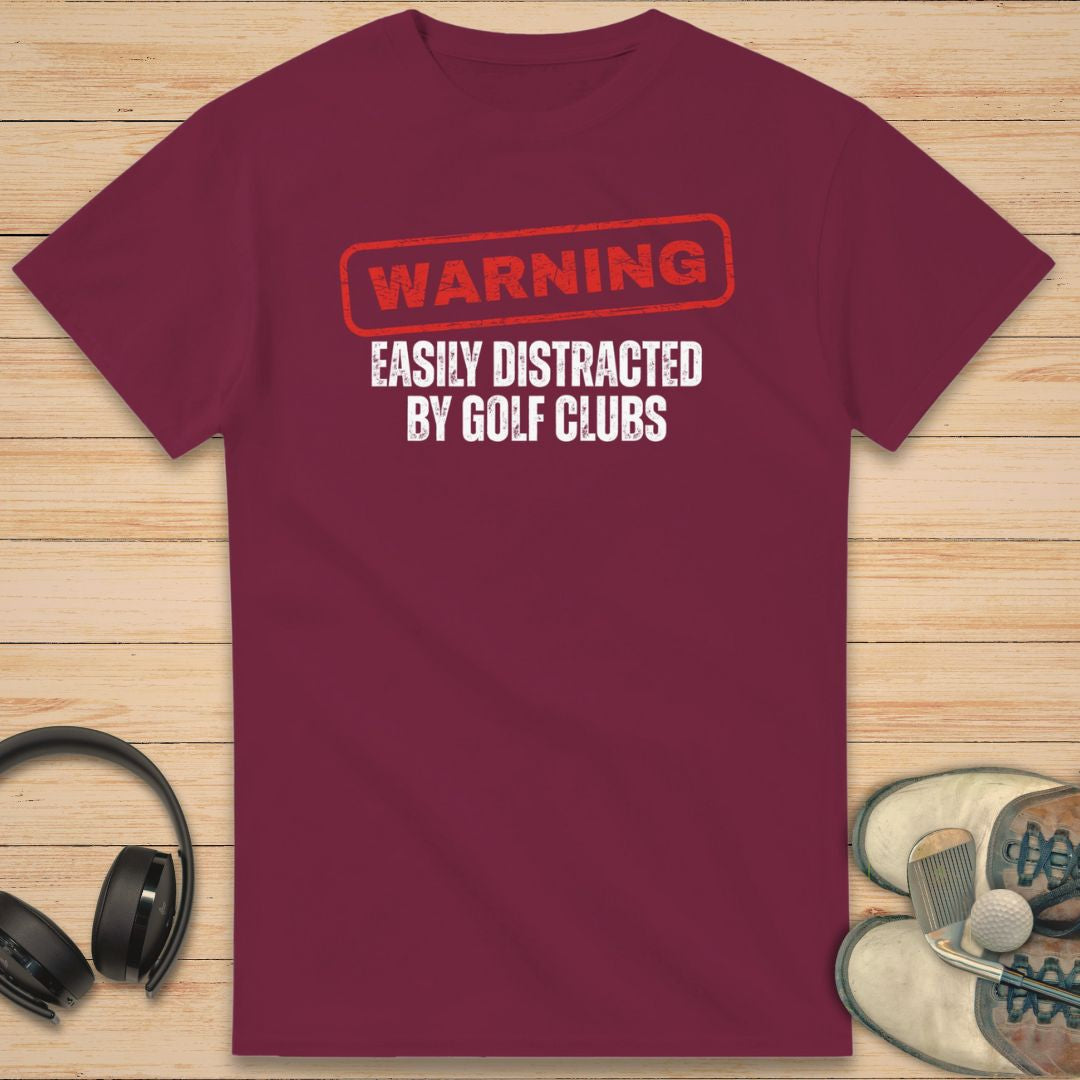 Warning T-Shirt in Maroon on a light wooden floor lying next to headphones and golf shoes.