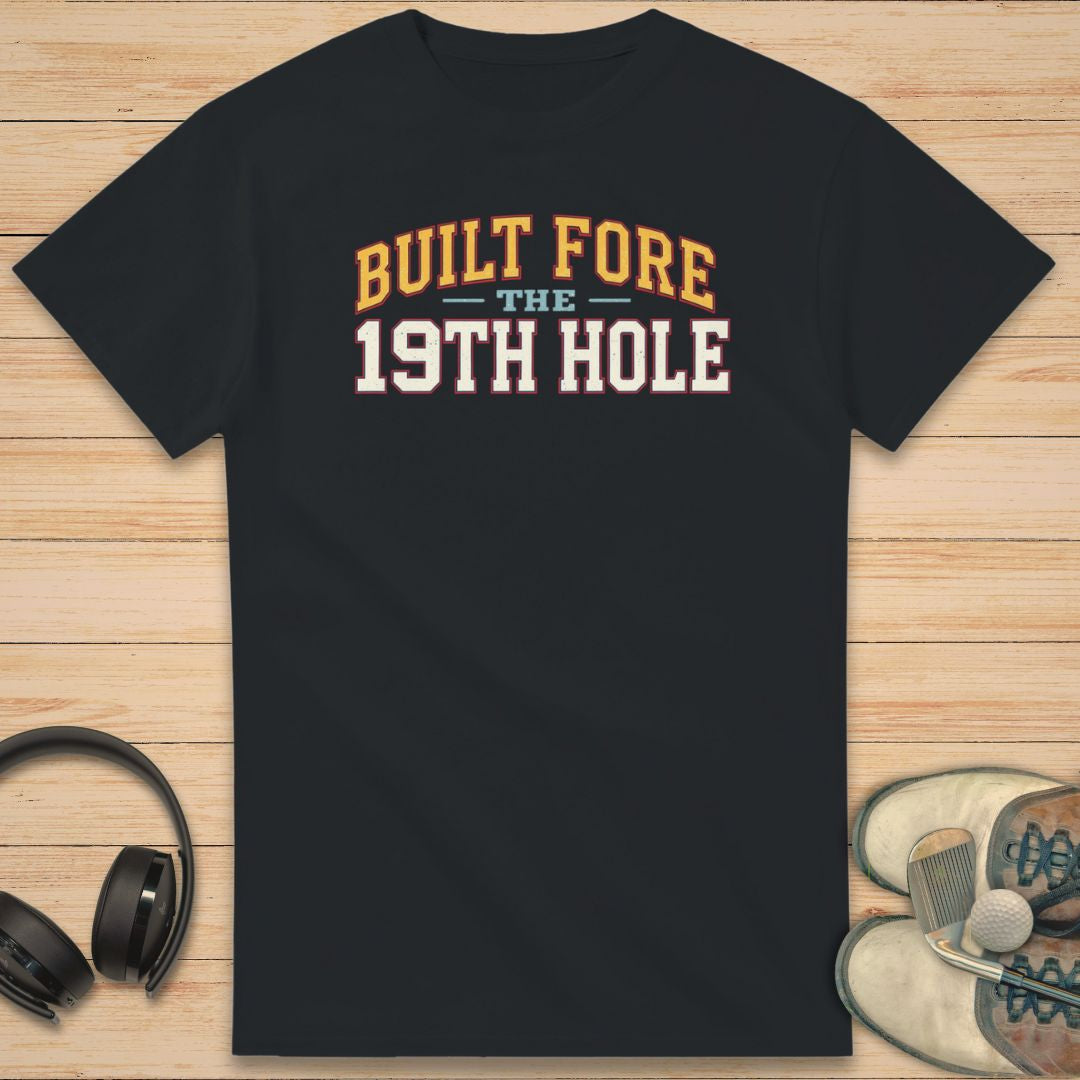 Built Fore T-Shirt in on a light wooden floor lying next to headphones and golf shoes.