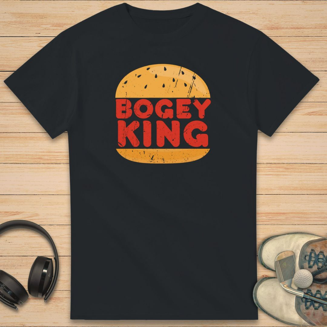 Bogey King T-Shirt in Black on a light wooden floor lying next to headphones and golf shoes.