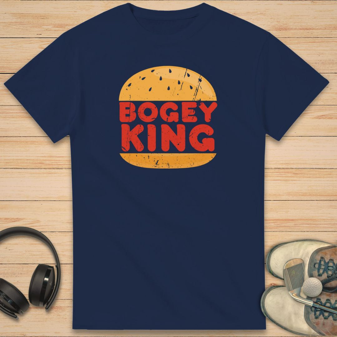 Bogey King T-Shirt in Navy on a light wooden floor lying next to headphones and golf shoes.