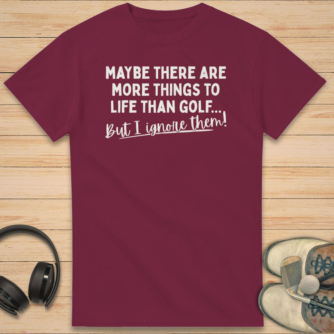 More Things T-Shirt in Maroon on a light wooden floor lying next to headphones and golf shoes.